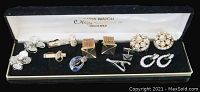 Vintage jewelry items including cufflinks, earrings, pins, and a tie clip displayed inside a black velvet-lined Oleg Cassini watch case.