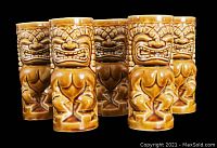 Photo showing set of five ceramic tiki tiki glasses arranged side-by-side, highlighting carved facial and body details with brown and tan glaze.
