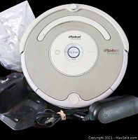 Photos show a white iRobot Roomba robotic vacuum with a central control panel and three buttons, accompanied by a black charging station dock, power adapter with cable, and a plastic bag containing user manual or spare parts. The Roomba shows signs of use and the lot is untested.