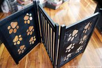 Black wooden pet gate with three panels including paw cutouts and vertical slats placed on hardwood floor.