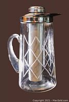 Front view of the clear glass pitcher showing the etched diamond design, curved handle, and shiny chrome lid with pour spout. Inside is the aluminum chill tube.