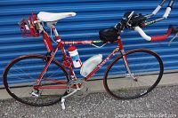 Front left side view of red Viner road bike leaning against blue storage container showing saddle, seatpost rack, water bottle and frame.