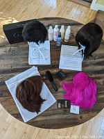 Top down view showing all four wigs on white wig stands, wigs include short dark pixie human hair, a shorter bob highlighted synthetic hair wig, longer dark synthetic bob wig, and vibrant pink synthetic bob wig with tags. Hair care products and combs arranged around.