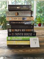 Stack of nine science fiction and fantasy paperback and trade paperback books on a wooden table with a window and plants in the background.