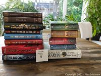 Full view of stacked hardcover books on a wooden table with green plants and chair in the background, showing titles such as Miss Peregrine's Home for Peculiar Children, The Hunger Games series by Suzanne Collins, The Secret Commonwealth and The Book of Dust by Philip Pullman, and multiple volumes of A Song of Ice and Fire by George R.R. Martin.