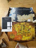 Photo shows a map poster from the Game of Thrones series, a colorful Lord of the Rings map poster, and a Sylvia Plath quote poster partially unrolled on a wooden floor.