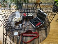 Black metal foldable pet crate with door open on wooden tabletop showing included accessories inside and around it: pet bowls on a raised holder, pet grooming kit in black case, paw clippers, Kong ball, and red dog collar.