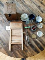 Wooden Rival washboard, hand mixer, metal container, enamelware cups, glass bottles, pipe, and teacups on wooden surface.
