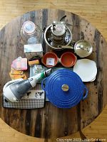 Overview of all kitchen items on wooden round table, including La Sera braiser, Black & Decker vacuum, containers, bowls, cutting boards, coasters