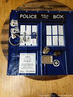 Blue plastic Dr. Who Tardis design shower curtain laid flat, showing white window panes, words 'Police Public Call Box' and 'Pull to Open'. Assorted metal and plastic bathroom accessories arranged on curtain.