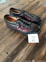 Pair of John Fluevog men's black leather shoes with red flame detail on sides, displayed on a wooden surface with auction card showing number 61.