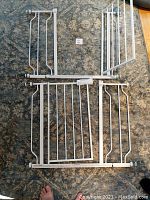 Overview of two white metal pressure-mounted baby gates with expansions laid on carpet. Adjustable width 29-39 inches.