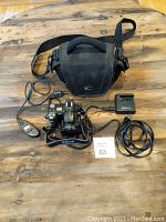 Full kit displayed on wooden table: Nikon camera body with attached neck strap, wired remote control, Nikon MH-24 battery charger with power cord, and black padded Nikon camera bag.
