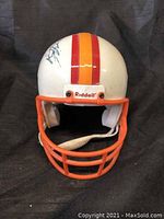 Front view of miniature helmet with signature on top, showing orange facemask and centered orange stripes.