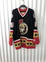 Front view of black Koho Ottawa Senators hockey jersey with Ottawa Senators logo, number 18 on sleeve, red and gold striping at bottom and sleeves.