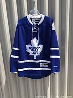 Front view of Toronto Maple Leafs blue hockey jersey hanging on white hanger. Shows logo, neckline, and sleeve stripes.