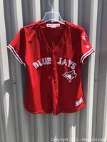 Front view of red Blue Jays baseball jersey showing 'BLUE JAYS' text and Blue Jays logo patch on front and buttons down center.