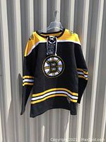 Front view of black Boston Bruins hockey jersey sweater with yellow and white stripes and lace-up collar, hanging on hanger.