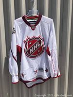 Front view of white NHL jersey showing large maroon and silver NHL shield logo on chest with maroon trim at collar, cuffs, and hem.