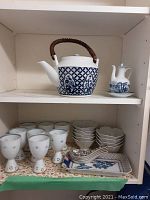 Upper shelf with blue and white teapot with woven handle, and smaller blue and white pourer. Lower shelf with 8 delicate glasses, 9 small white floral bowls, and ceramic spoons on a floral tray.