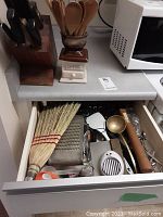 Wooden knife block with multiple knives, ceramic holder with wooden spoons, drawer with kitchen brooms, metal grater, brass ladle, and other kitchen tools.
