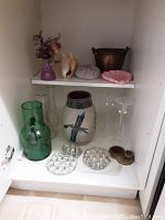 Shelf with large green glass vase, smaller clear vase, flower frogs, ceramic pot with bird design, and glass candlesticks.
