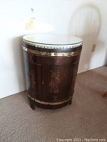Front view of the round carved drum table with figures and decorative motifs, showing the glass top and brass nail accents.