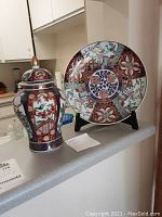 Photo shows the jar with lid and decorative plate side by side on a kitchen counter, displaying the overall size and ornate decorated surfaces of each piece.