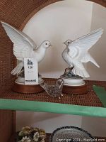 Two white ceramic doves on wooden stands and small glass bird figurine on wicker shelf.