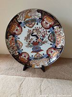 Front view of large painted ceramic dish on stand showing Imari floral and geometric designs in red, blue, green, and gold.