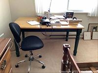 Rectangular maple topped table with dark painted turned legs alongside black cushioned office chair with chrome base wheels in room with carpet flooring and window.