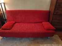 Red foldable sleeper sofa in upright position with two cushions on a rug near wooden furniture.