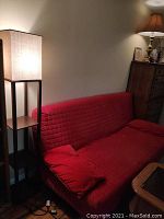 Floor lamp standing beside a red couch and a cabinet, with a lit square fabric lampshade and dark wooden frame.