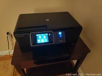 Front and touchscreen interface view of black HP Photosmart Plus wireless printer on wooden table, powered on and showing main menu.