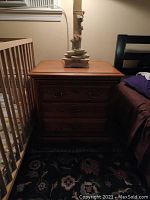 Front view of solid wood nightstand with two drawers and metal handles, on patterned carpet near crib and bed.