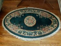 Top view of oval area rug showing central floral medallion and surrounding floral border on green background with fringe edges laid on wooden floor.