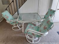 View of composite square patio table with glass top surrounded by three cushioned armchairs with floral print cushions and umbrella installed