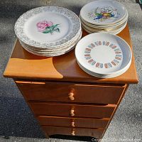 Photo of small solid wood 4-drawer cabinet with stacks of assorted decorative plates on top.