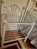 Side angle view of white enamel metal end table with glass top and decorative finials, placed next to a bed.