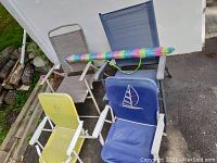 Four portable beach chairs of various colors and designs plus a rolled colorful beach mat resting on them, placed outdoors on concrete near a white wall and wooden logs.
