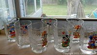Photo showing eight assorted Disney-themed drinking glasses on a windowsill with clear glass and colorful character decals.