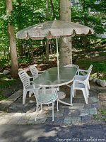 Full view of patio set showing glass table, umbrella, and six chairs on a stone patio surrounded by trees.