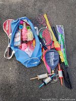 Blue Wilson sports bag open with tennis rackets, badminton rackets, kite strings spools, kite parts and volleyball net poles on pavement.
