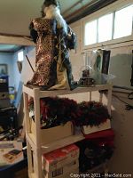 White shelving rack holding a tall Santa Claus figurine, Christmas wreaths in boxes, and a snow globe ornament near a window