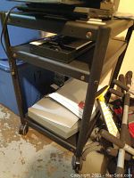 Black metal cart on wheels with shelves holding multiple electronics and devices including a Cisco router and switch.