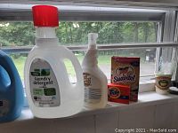 Various cleaning liquids including a white plastic jug labeled laundry detergent with a red cap, a white-spray bottle, a pack of Suavitel sheets, and a Tide detergent box on a windowsill