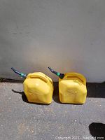 Photo showing two yellow plastic diesel fuel cans with attached nozzles placed on the ground against a wall.
