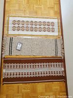 Three table runners laid flat on wood floor. Top is a cream runner with brown and yellow diamond patterned stripes, middle is thick natural wool runner with black stripes and fringe, bottom is brown runner with white floral geometric design.