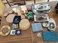 Lot of assorted serving and dinnerware items on wooden table including cutting boards, woven trivets, silver plated trays, flatware sets in cases, and serving utensils.