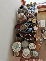 Photo showing a view of multiple large and small vintage ceramics including bowls, vases, mugs, and a ceramic fish plaque arranged on tables and a window ledge